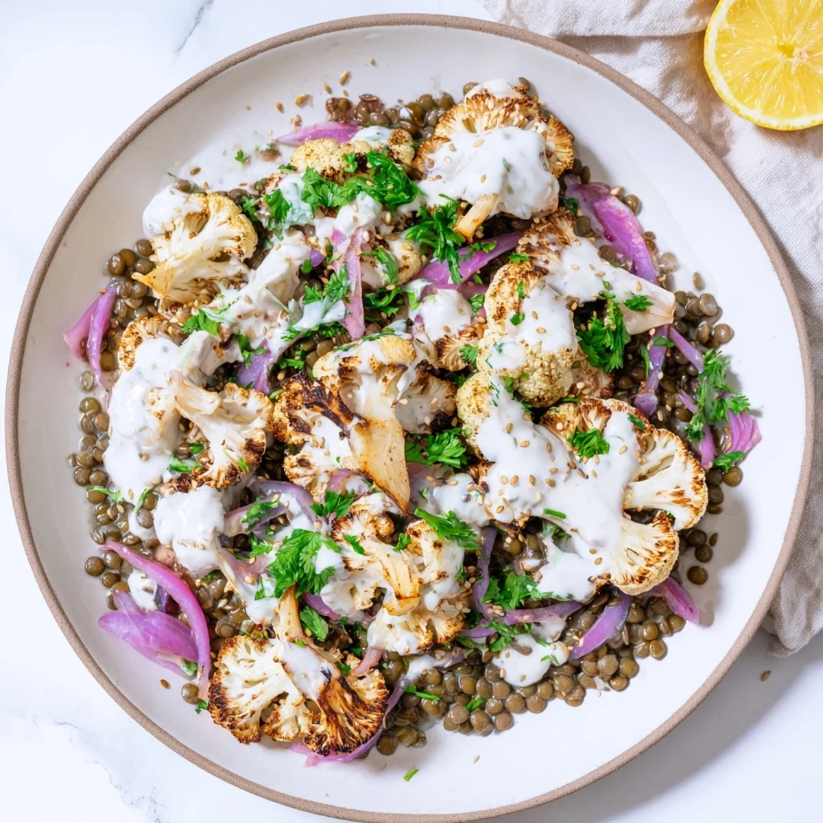 Golden roasted cauliflower florets seasoned with cumin and paprika are arranged over a bed of zesty lemon lentils, garnished with fresh herbs and toasted sesame seeds, ready to be drizzled with creamy tahini yogurt.